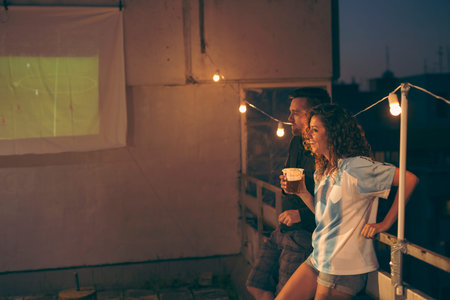 Couple of football fans standing on a building rooftop terrace, hanging out and drinking beer while...