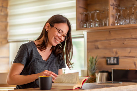 Beautiful young woman sitting at kitchen counter in the morning, drinking coffee and reading a book,...