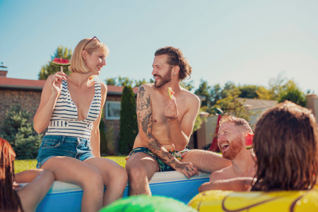 Group of cheerful young friends sitting at the edge of a swimming pool, sunbathing and making a...