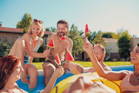 Group of cheerful young friends sitting at the edge of a swimming pool, sunbathing and making a...