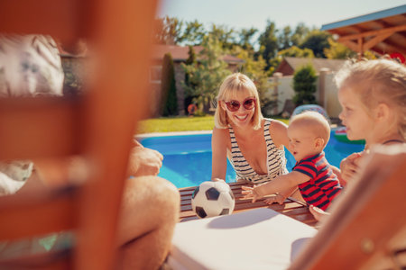 Young parents having fun playing with their children on a sun bed by the swimming pool, relaxing...