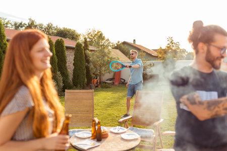 Group of cheerful young friends having a backyard barbecue party, grilling meat, drinking beer,...