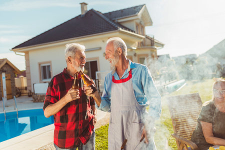 Two senior men drinking beer while grilling meat at backyard barbecue with their wifes sitting at...