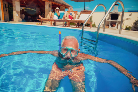 Group of senior friends relaxing and enjoying summer vacation, elderly man having fun snorkeling in the pool while women are sunbathing on sun beds by the poolの写真素材