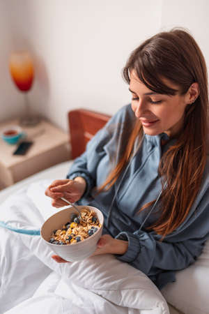 Beautiful young woman wearing pajamas sitting in bed in the morning, pouring milk into a bowl of cereal with fresh berries for breakfast and enjoying free time at homeの写真素材
