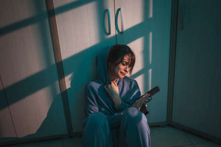 Upset woman sitting on the floor in the dark, holding a gun and crying after committing murder in self-defense; scared woman victim of domestic violence holding a gun for self-defenseの写真素材