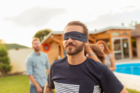 Group of cheerful young friends having fun at poolside summertime outdoor party, playing blind man's...