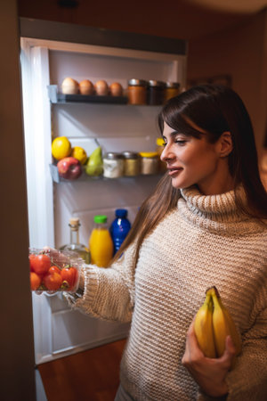 Beautiful young woman standing next to an opened fridge in the kitchen at night, enjoying eating...