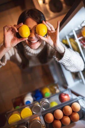 Beautiful young woman standing next to an opened fridge in the kitchen at night, enjoying eating pastaの写真素材