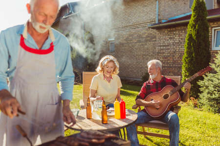 Senior neighbors having fun spending sunny summer day together outdoors, having a backyard barbecue party, playing the guitar and singingの写真素材