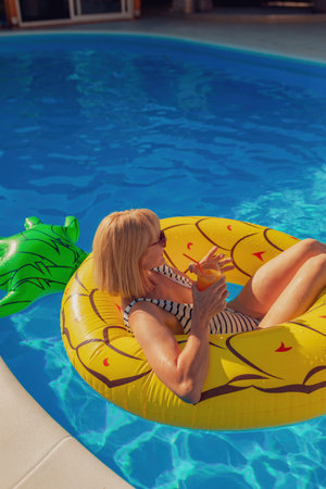 Attractive young woman wearing swimsuit drinking cocktail while floating on pool raft, enjoying...