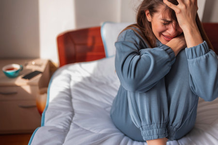 Frustrated young woman wearing pajamas, sitting on bed in the morning, wiping tears using paper...
