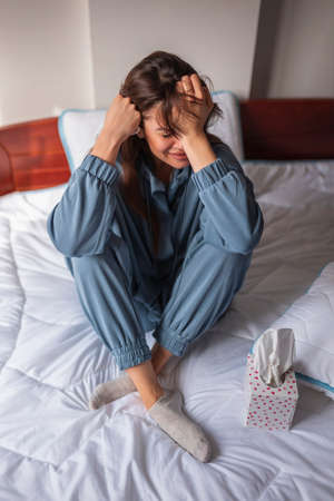 Frustrated young woman wearing pajamas, sitting on bed in the morning, wiping tears using paper tissue while sobbingの写真素材