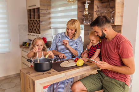 Cheerful young parents having fun in the kitchen with their children, sitting at kitchen counter, cutting and eating vegetablesの写真素材
