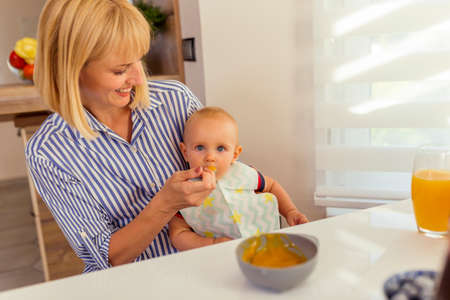 Beautiful young mother sitting at dinning room table, holding her baby boy in her lap and feeding him with a fruit porridgeの写真素材