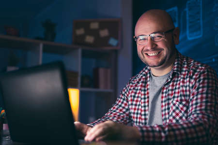 Man working overtime in home office sitting at his desk, having a video call using laptop computerの写真素材