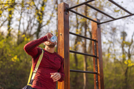 Athletic young man carrying gym bag, drinking water after an intense outdoor workout in a street...