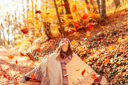 Beautiful young woman spending sunny autumn day in nature, sitting on a bench in the forest, smiling while hiding and peeking behind the bookの写真素材
