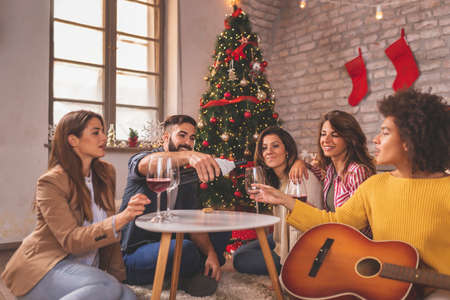 Group of friends sitting by nicely decorated Christmas tree, celebrating Christmas at home, playing the guitar, drinking wine and singing Christmas songsの写真素材