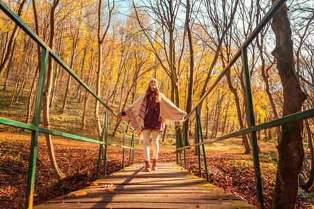 Woman walking down the bridge over a stream on the forest path covered with colorful fallen leaves, enjoying spending sunny autumn day outdoors in natureの写真素材