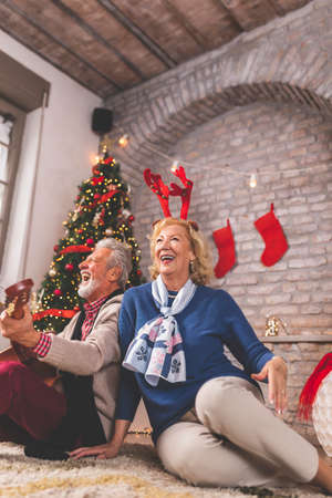 Elderly couple having fun celebrating Christmas at home, sitting by nicely decorated Christmas tree, drinking coffee and looking through old photo albumsの写真素材