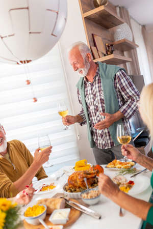 Two senior couples having fun while celebrating Thanksgiving together at home over traditional dinner, pouring wine and making a toastの写真素材