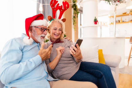 Senior couple sitting on the sofa by nicely decorated Christmas tree, celebrating Christmas at home, having fun while drinking coffee and eating gingerbread cookiesの写真素材