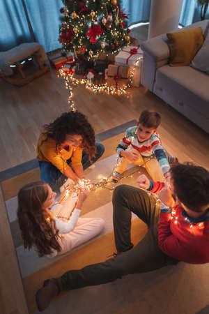 Parents celebrating New Years Eve at home with kids, sitting by the Christmas tree, holding illuminative numbers 2022 representing the upcoming New Yearの写真素材