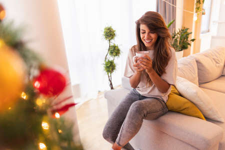 Beautiful young woman wearing costume reindeer antlers having fun peeking through candy cane heart while decorating Christmas treeの写真素材