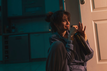 Depressed drunk woman sitting on the floor in the dark with smeared makeup, drinking wine from a...