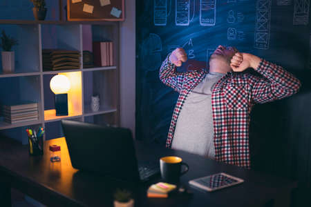 Man sitting at his desk in home office, drinking coffee and using laptop computer while working lateの写真素材