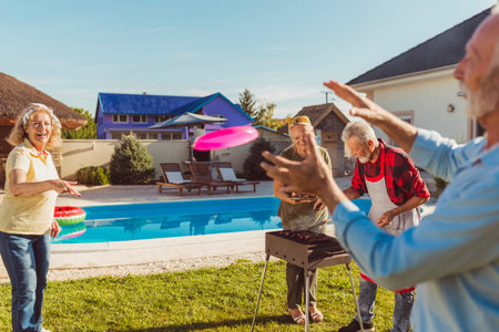 Group of cheerful senior friends eating lunch in the backyard by the pool, gathered around the...