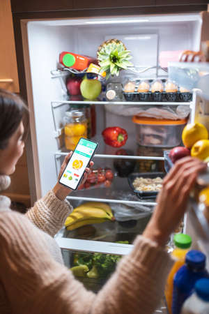 High angle view of a beautiful young woman standing next to an opened refrigerator door, ordering fresh fruit and vegetables online for home delivery using a smart phone appの写真素材