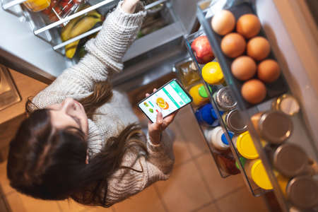 High angle view of a beautiful young woman standing next to an opened refrigerator door, ordering fresh fruit and vegetables online for home delivery using a smart phone appの写真素材