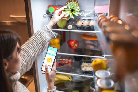 High angle view of a beautiful young woman standing next to an opened refrigerator door, ordering fresh fruit and vegetables online for home delivery using a smart phone appの写真素材