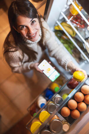 High angle view of a beautiful young woman standing next to an opened refrigerator door, ordering fresh fruit and vegetables online for home delivery using a smart phone appの写真素材