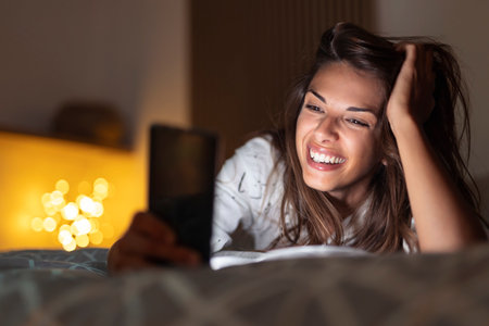 Beautiful woman relaxing at home late at night, sitting in bed having video call using tablet computerの写真素材