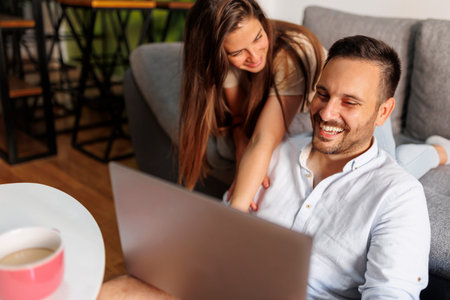Couple in love enjoying leisure time at home, drinking take away coffee and surfing the Internet using laptop computer and smart phoneの写真素材