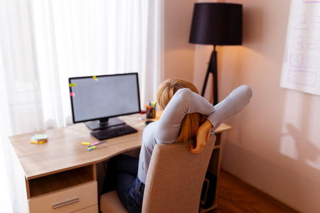 Woman stretching out while sitting at her desk in home office, tired while working overtimeの写真素材
