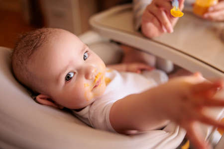 Mother feeding her baby boy using spoon, introducing first solid food meal, baby sitting in high chair all messy and staied with porridge, looking towards the cameraの写真素材