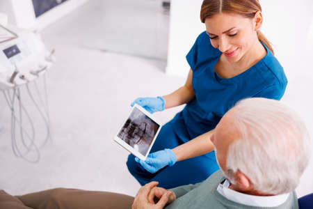 Doctor showing jaw x-ray on tablet computer to patient sitting in dental chair at dentist officeの写真素材