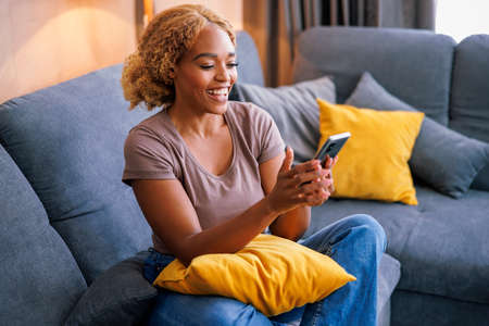 Beautiful young woman relaxing at home, sitting on living room couch and smiling while having video call using smart phoneの写真素材