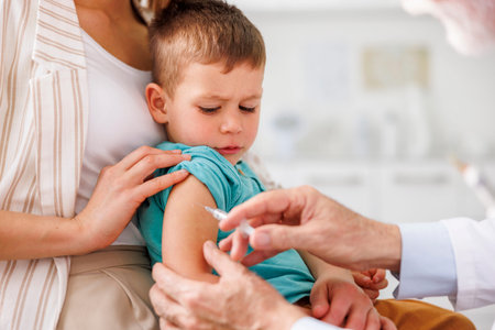 Cute little boy sitting in mother's lap getting vaccinated by doctor at hospital, doctor...