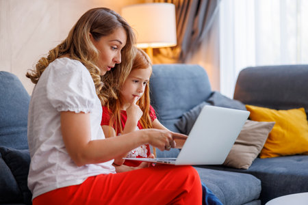 Beautiful mother helping her little daughter with school project, folding and cutting paper using scissorsの写真素材
