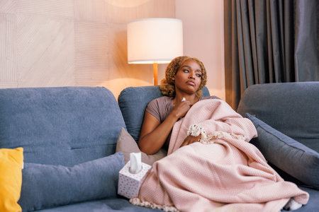 Woman lying sick at home covered with blanket on living room couch, suffering in pain having fever and fluの写真素材