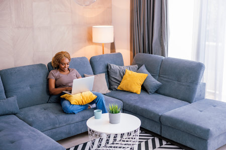 Female freelancer sitting on living room couch working from home using laptop computer and drinking coffee; woman working remotely from homeの写真素材