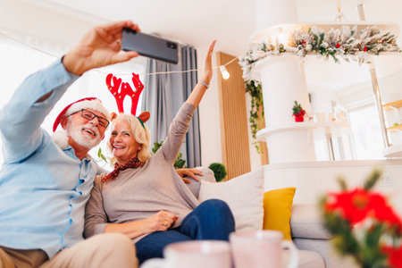 Happy senior couple sitting on the living room sofa celebrating Christmas at home, having fun taking selfies using smart phoneの写真素材