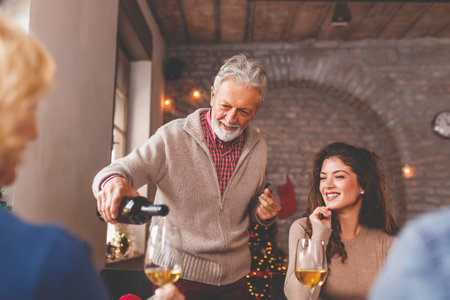 Family having a Christmas dinner at home, enjoying their time together, talking to each other and laughing while senior man is pouring wineの写真素材