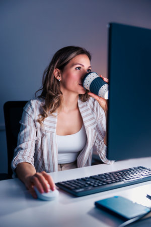 Female web designer working late in the office, drinking coffee and using laptop computer; casual business woman working overtimeの写真素材