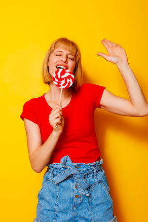 Beautiful blond woman in trendy summer wear holding candy as mike singing and dancing on yellow color backgroundの写真素材